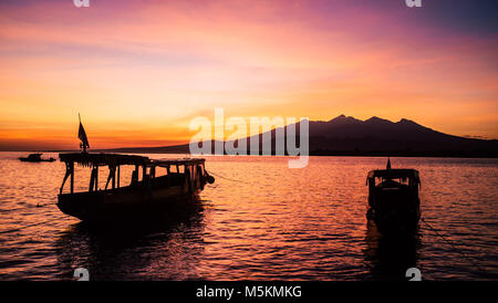 The sunrise above the volcano in Bali from the Gili Islands can be seen with two boats in the foreground Stock Photo