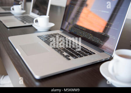 Multiple computers with coffee in a office environment close up showing lines of code on screen from side. Stock Photo