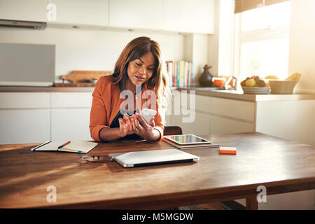 Smiling young African female entrepreneur reading text messages while sitting at her kitchen table working from home Stock Photo
