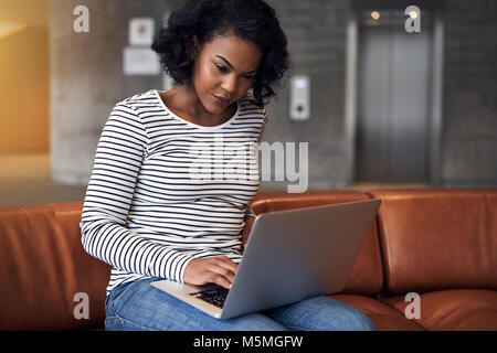 Focused young African college student sitting alone on a couch on campus studying online with a laptop Stock Photo