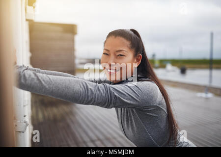 Smiling young Asian woman in sportswear stretching against a wall outside before going for a jog Stock Photo