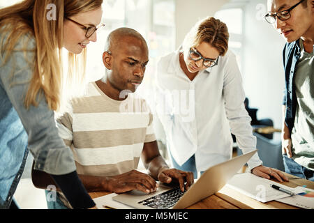 Diverse group of focused young work colleagues standing together at an office table using a laptop Stock Photo