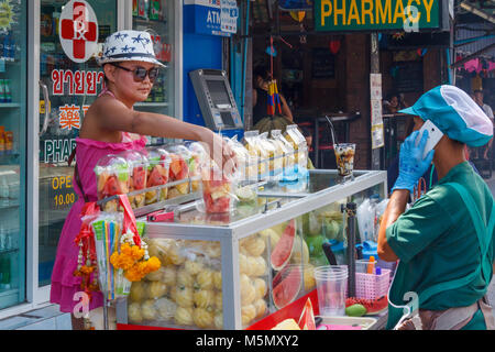 Woman in hat and sunglasses buying fruit from a street vendor on mobile phone, Bangla road, Patong Beach, Phuket, Thailand Stock Photo