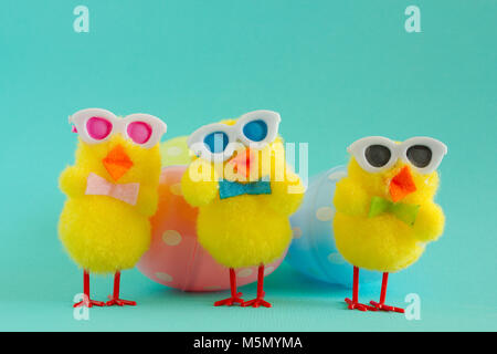 Three groovy yellow fuzzy pom pom Easter chicks sporting sunglasses for a cool look. Three eggs, one pink, one yellow & one blue, all with white spots Stock Photo