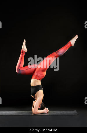Athletic woman doing a headstand with bow and arrow legs as she works out on a yoga mat in a side view over black with copy space Stock Photo