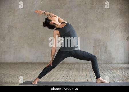 Young woman practicing yoga, doing Reverse Warrior exercise, Viparita Virabhadrasana pose, working out, wearing sportswear, black pants and top, indoo Stock Photo