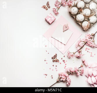 Easter background with flat lay of eggs,gifts, decorations blossom and blank greeting card mock up on white desktop, top view, pink pastel color Stock Photo