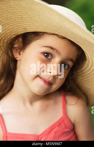 Portrait of smiling beautiful toddler in wide-brimmed hat at green of summer park. Stock Photo