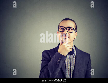 Young happy man in suit and glasses rubbing chin and daydreaming looking away on gray. Stock Photo