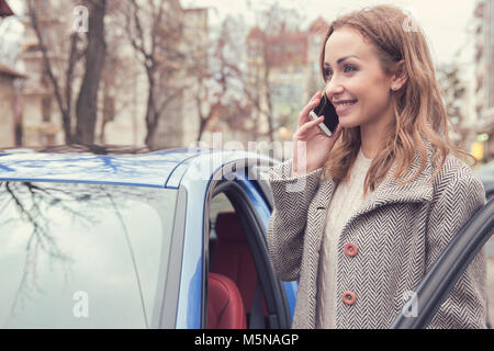 Pretty woman in outerwear having phone call standing on street near automobile and smiling. Stock Photo