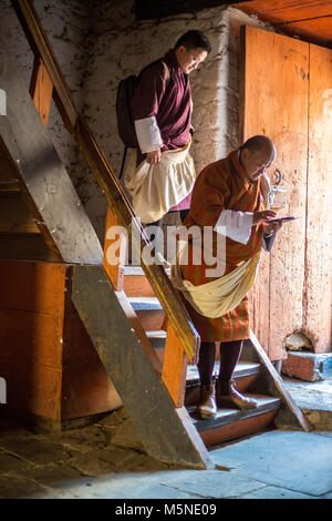 Jakar, Bhutan.  Men in Traditional Male Attire (Gho) Descending Stairway inside the Jakar Dzong (Monastery), One Using his Cell Phone. Stock Photo