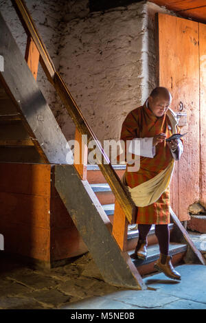 Jakar, Bhutan.  Man in Traditional Male Attire (Gho) Descending Stairway inside the Jakar Dzong (Monastery), Using his Cell Phone. Stock Photo