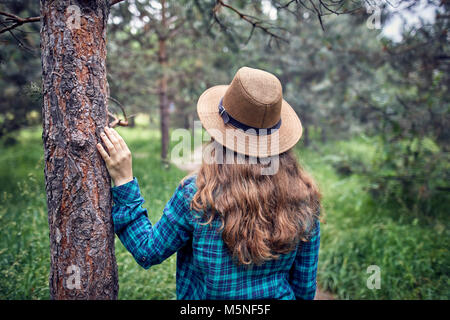 Young woman in brown hat and green checked shirt with long hair touching the pine tree in the forest