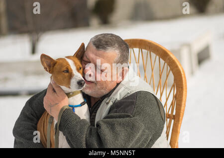 Portrait of Caucasian senior man with his cute basenji dog sitting in wicker chair at sunny winter day Stock Photo