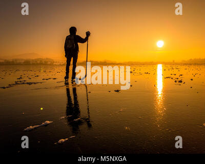 Scenic panoramic view of the silhouette of a young hockey player standing on a frozen lake with amazing reflections in beautiful golden evening light  Stock Photo