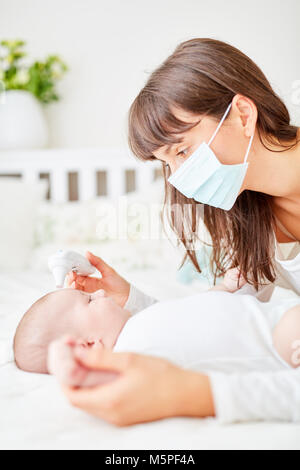 Woman as a doctor or nurse with surgical mask measures fever in infant Stock Photo