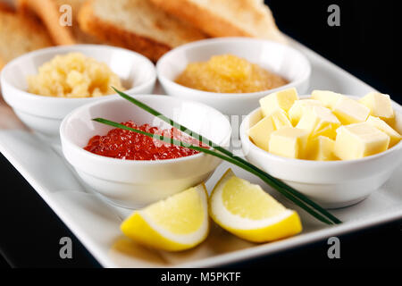 On a white plate of assorted with red white and pike caviar served baguette and butter. The bowl is decorated with lemon and shallots. Background blac Stock Photo