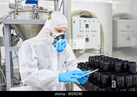 Confident young worker wearing coverall and respirator using digital tablet while taking inventory in warehouse of pharmaceutical factory Stock Photo