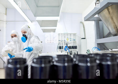 Team of confident factory workers wearing rubber gloves and coveralls using digital tablet in order to take notes while taking inventory in sport nutr Stock Photo