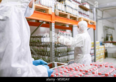 Working process in food factory warehouse: group of workers wearing coveralls and safety masks wrapped up in loading goods, one of them holding digita Stock Photo