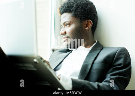 Waist-up portrait of pensive African American businessman wearing stylish suit sitting on window sill and enjoying picturesque view, he holding laptop Stock Photo