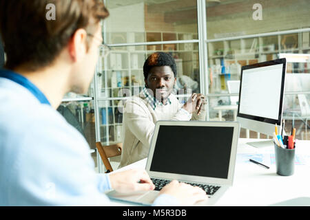 Handsome African American manager sitting at desk and chatting with his male colleague while taking short break from work, interior of open plan offic Stock Photo