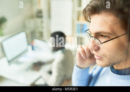 Close-up shot of confident young manager in eyeglasses standing at modern open plan office and talking to his client on smartphone, view through panor Stock Photo