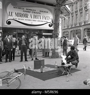 1958, historical, Paris, France. Infront of a cinema showing the french drama film, ' Le Miroir A Deux Faces', a small crowd stand watching some street theatre or magic, as a performer or bucker, who has laid his props out on the pavement, lights a fire in preparation for his act. Stock Photo