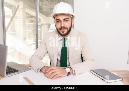 Portrait of handsome young engineer wearing white hardhat sitting at table with work documents and looking at camera, copy space Stock Photo