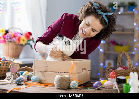 Portrait of young woman smiling happily while playing with cute Easter bunny sitting in wooden box Stock Photo