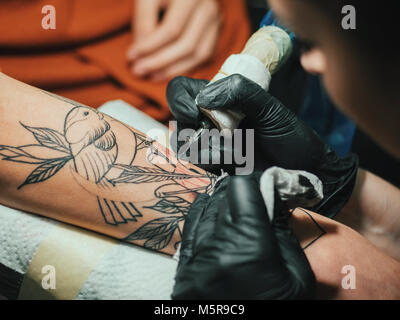 Tattoo artist at work. Woman in black latex glove tattooing a young man's hand with colorful picture in studio. Close up. Stock Photo
