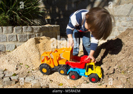 little three year old boy playing in the sand with a digger and dump truck. Stock Photo