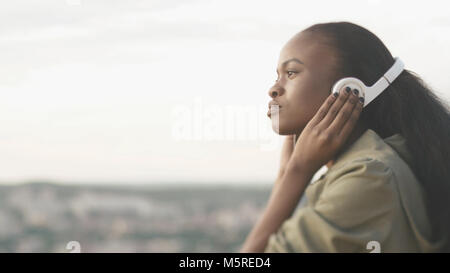 Side portrait of the beautiful attractive young african girl with natural make-up listening to music in headphones at the blurred background of the city. Stock Photo