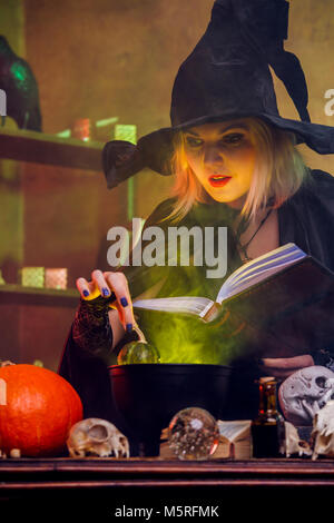 Picture of witch blonde in black hat with book brewing potion in pot Stock Photo