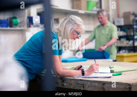 Senior businesswoman is doing paperwork in her stock warehouse. Her husband is in the background measuring fabric. Stock Photo