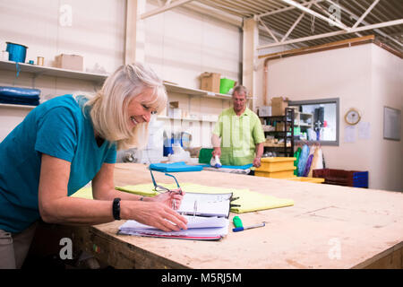 Senior businesswoman is doing paperwork in her stock warehouse. Her husband is in the background ironing fabric. Stock Photo