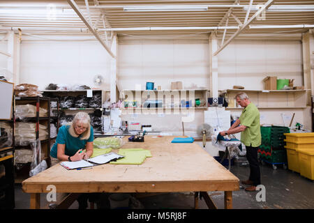 Senior businesswoman is doing paperwork in her stock warehouse. Her husband is in the background ironing fabric. Stock Photo