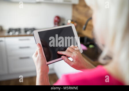 Back portrait of senior lady standing at kitchen using tablet Stock Photo