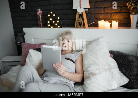 Portrait of senior lady lying on bed reading ebook Stock Photo