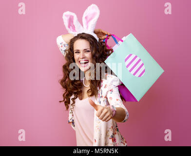 Festive bunny and eggs season. happy modern woman in Easter bunny ears isolated on pink with Easter shopping bag showing thumbs up Stock Photo