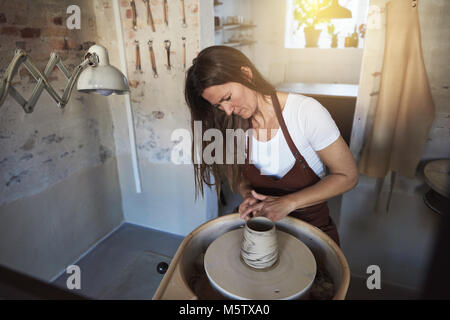 Artisan using a tool to creatively sculpt a piece of clay turning on a pottery wheel while working in her ceramic workshop Stock Photo