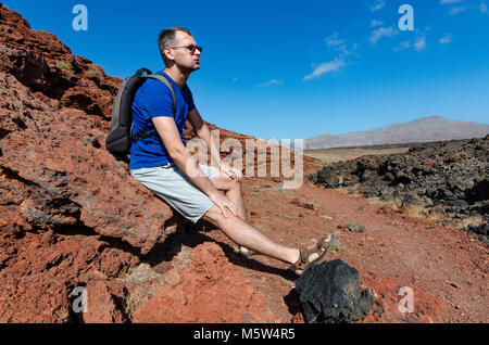 A man with a backpack sitting on a red rock and resting while trekking in volcanic landscape of Timanfaya National Park on Lanzarote, the Canary Islan Stock Photo