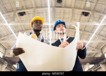 Bearded businessman wearing hardhat and suit studying blueprint with help of African American industrial engineer while carrying out inspection at pro Stock Photo