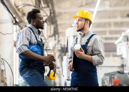 Multi-ethnic team of technicians wearing overalls chatting animatedly with each other while taking coffee break, interior of spacious production depar Stock Photo