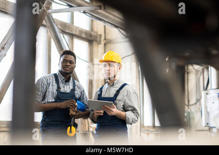 Multi-ethnic group of machine operators wearing overalls looking away pensively while taking coffee break in spacious production department of modern  Stock Photo