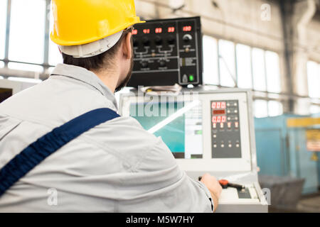 Back view of bearded operator wearing protective helmet focused on work while sitting in front of CNC machine, interior of production department on ba Stock Photo