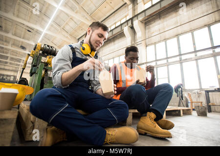 Multi-ethnic team of machine operators wearing overalls sitting at spacious production department of modern plant and eating instant noodles with chop Stock Photo