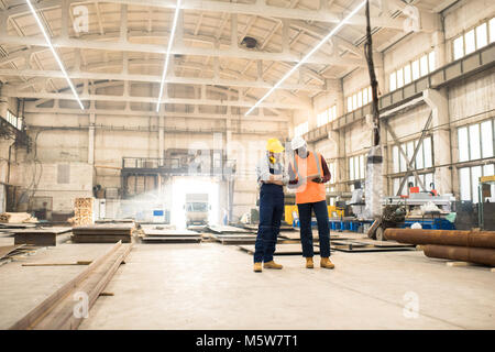 African American engineer wearing reflective vest and hardhat using digital tablet while discussing joint work with his colleague, interior of spaciou Stock Photo