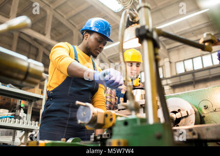 Multi-ethnic team of workers wearing overalls and protective helmets using lathe in order to machine workpiece, interior of spacious production depart