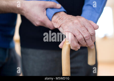 Senior Man's Hands On Walking Stick With Care Worker In Background Stock Photo
